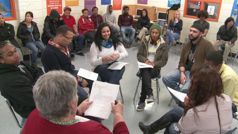 A multiracial group of people sit in a circle at a restorative justice training. More people sit in chairs lining the walls around the inner circle.