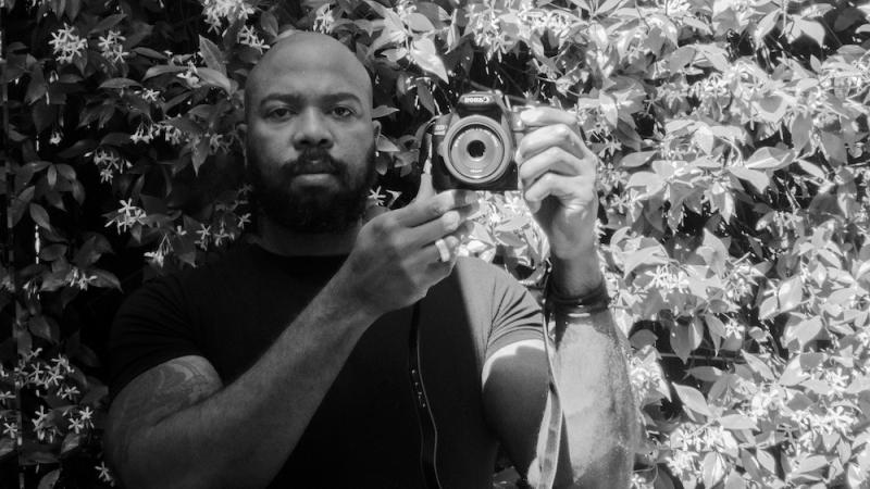 In this black and white photo, African-American filmmaker Daryl P Jones stands in front of flowering jasmine, looking directly at the camera. He holds up a digital video camera near his shoulder. He’s bald and has a thick beard.