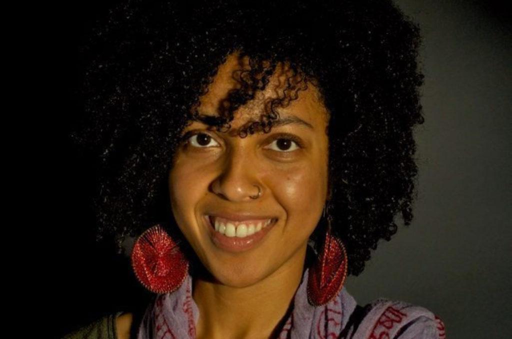 A headshot of New Day Filmmaker Najma Nuriddin, an African-American woman with wavy shoulder length hair and large red earrings. She looks directly at the camera and smiles widely.