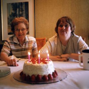 Dona and her mother, Mimi, sit at the dining room table, smiling for the camera. On the table, a white-frosted cake lined with strawberries with lit pink candles on top.