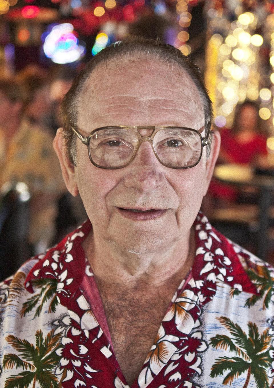 A still from the New Day Film Before You Know It. An balding, elderly man with glasses and a brightly patterned button down shirt looks into the camera with an amused grin. People and bright colored lights fill the blurred background.