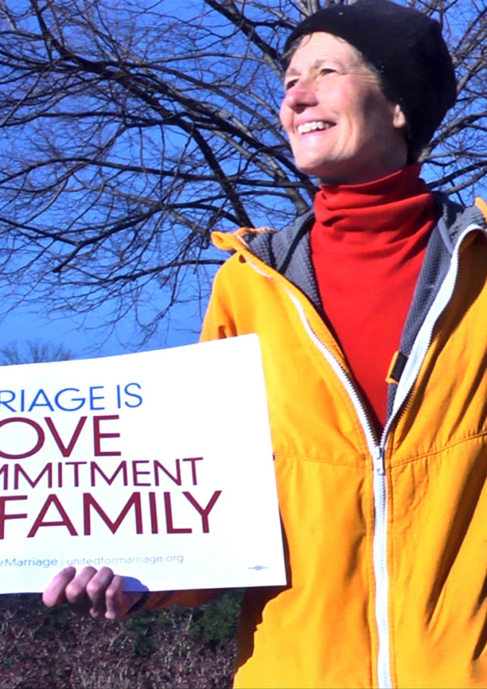 A white woman stands outside in front of a bare tree with blue sky. She smiles broadly, looking to the side. She is holding a white sign that reads “Marriage is Love, Commitment, Family” in blue and purple text. She wears a bright yellow zip up jacket which is half-unzipped to reveal a bright red turtleneck underneath and a black beanie on her head.
