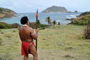 A Carib dressed in Carib garb is standing with a spear looking out at the ocean. 