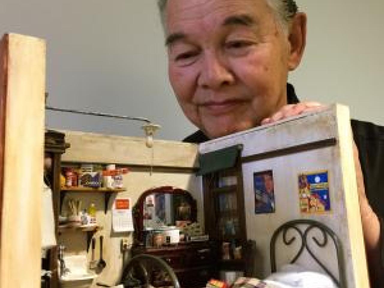 Frank Wong, an elder Chinese-American man, smiles as he peers into an intricately decorated miniature bedroom he made, complete with bed, vanity with mirror, art on the walls, and a sink.