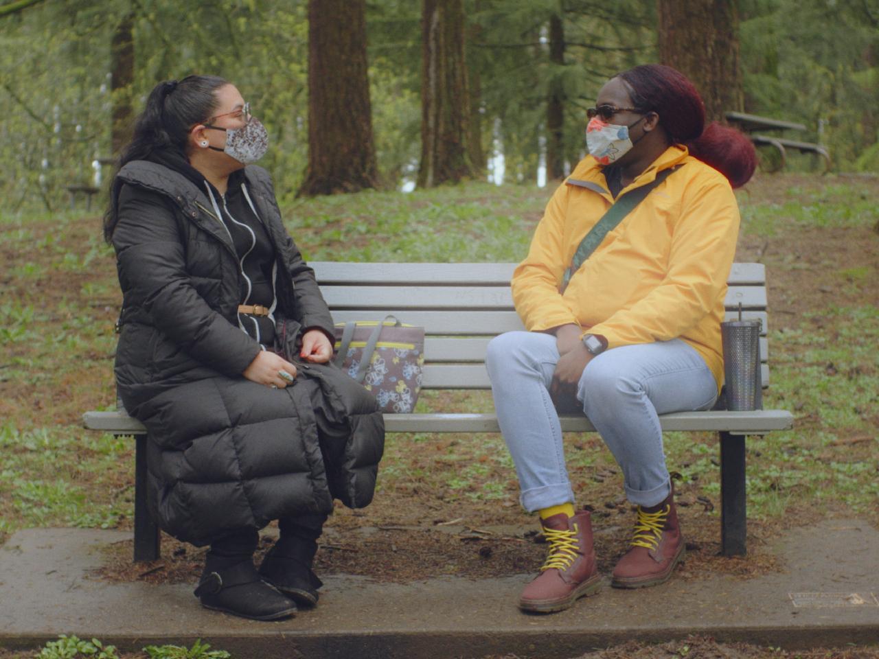 Two trans people sit on a park bench looking at one another. They wear cloth masks.