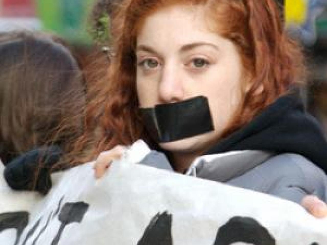 A young girl with red hair stares boldly into the camera in a protest. She has a large black strip of duct tape covering her mouth and she is holding up a protest banner. The other people holding up the banner are blurred in the background.