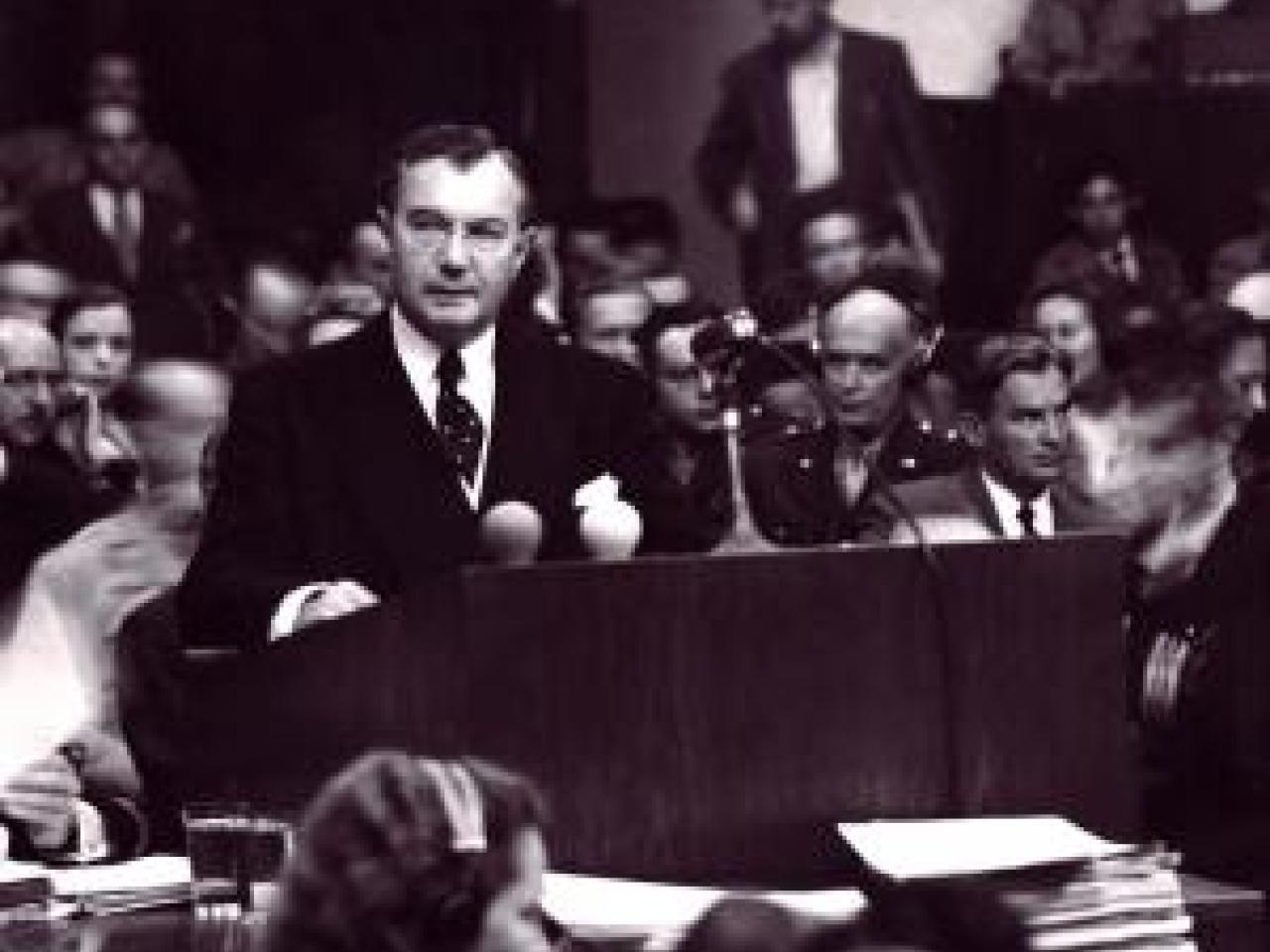 A black and white image from the film Nuremberg: Its Lesson for Today. A man in a suit stands at a podium with a serious expression looking slightly off to the side. Behind him sits a full audience of people, looking in different directions with serious expressions on their faces. In front of the podium sit women with headsets on and large stacks of paper in front of them.
