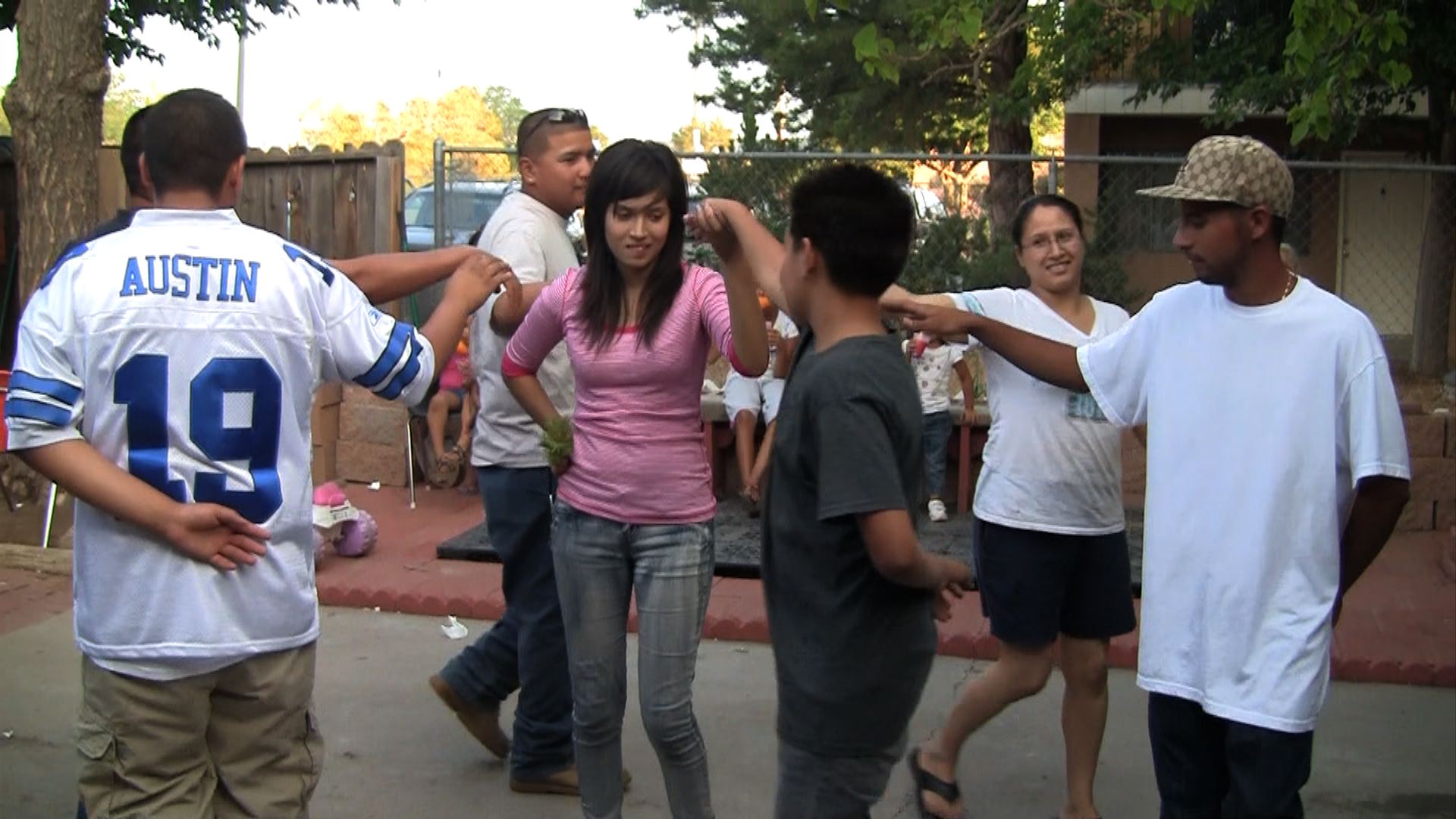 A group of seven people, mostly young, are dancing. They hold one arm outstretched and the other behind their backs. In the center, a girl and a boy are holding hands.