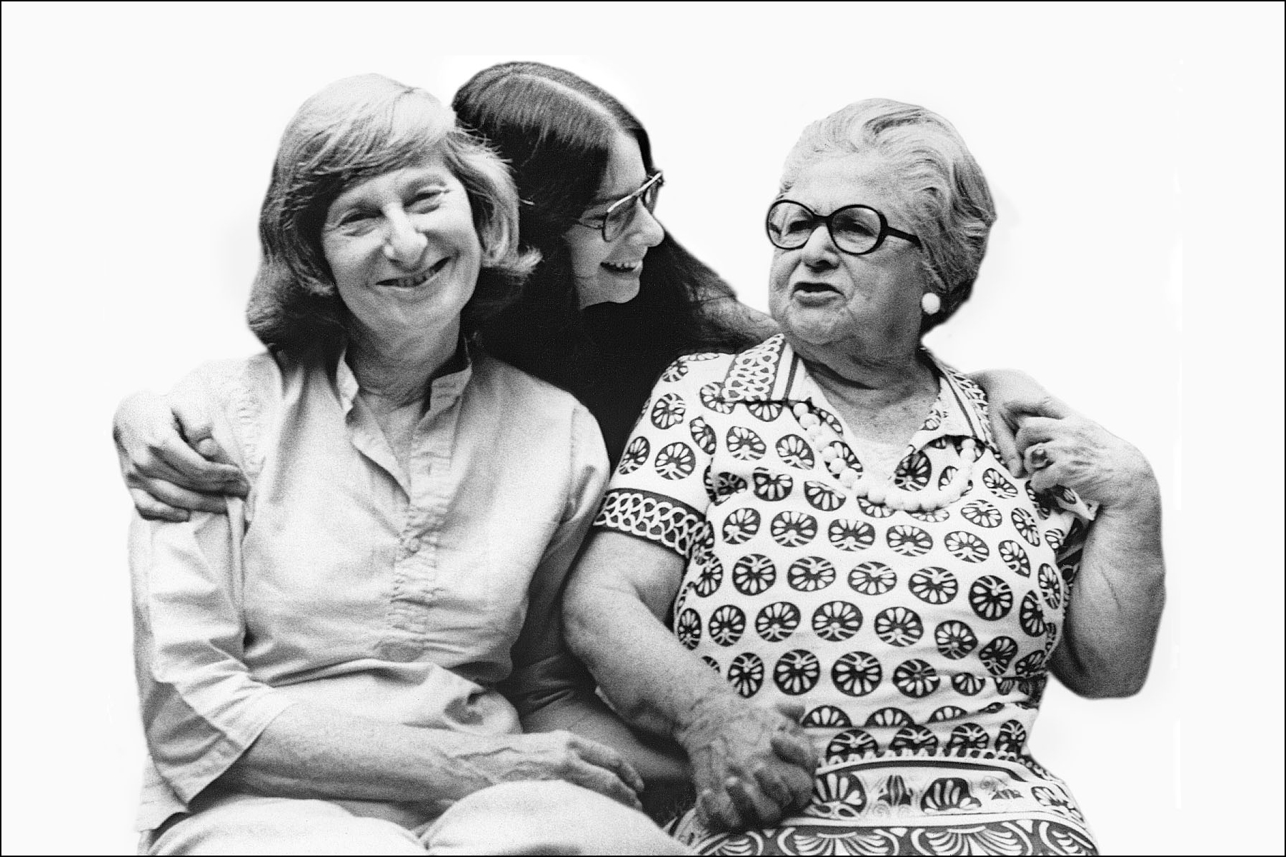 A black and white still from the New Day film, Nana, Mom and Me. A young woman stands behind her mother and grandmother with her arms around them. She glances affectionately towards her grandmother. The grandmother holds her granddaughter’s hand on her shoulder and her daughter's hand on her lap. Beside her, the mother smiles towards the camera.