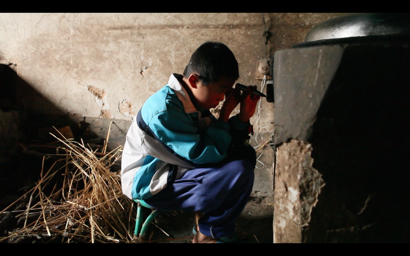 A young Chinese boy, maybe ten years old, sits on a low stool, working intensively on an electrical device attached to an old decaying structure.