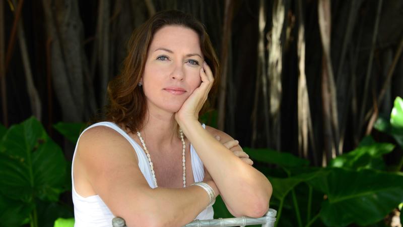 A woman with long brown hair and blue eyes rests on a chair and looks at the camera. 