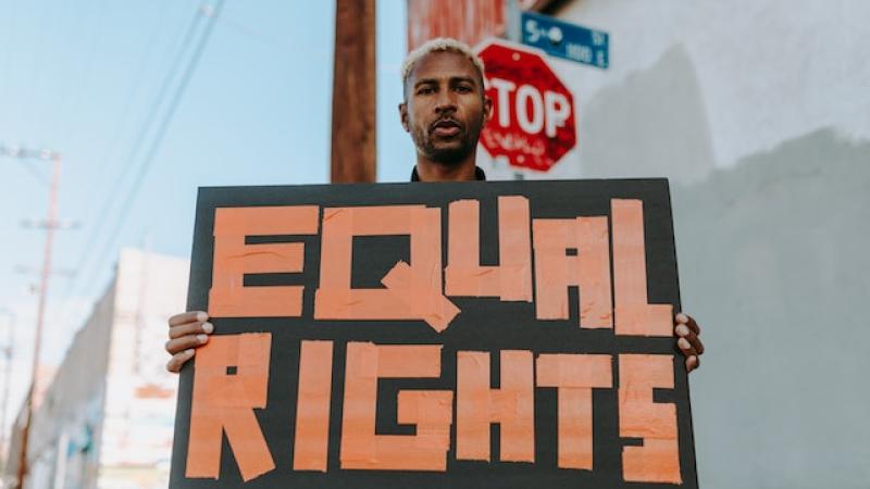 A man is holding up a homemade sign that says "equal rights". There is a stop sign and a cityscape behind him.