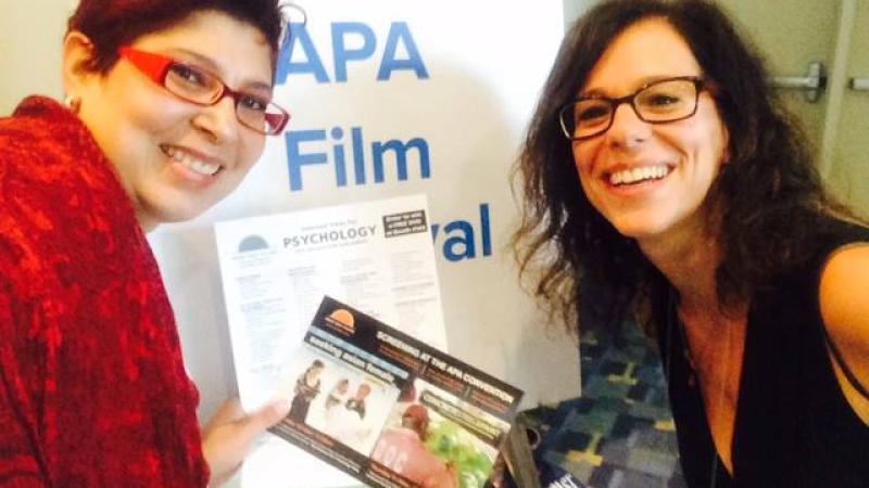 New Day Filmmaker Cindy Burstein and Gigi, from the APA social media, smile and lean into the frame in front of a board that reads “APA Film Festival.” Gigi holds a promotional New Day flyer.