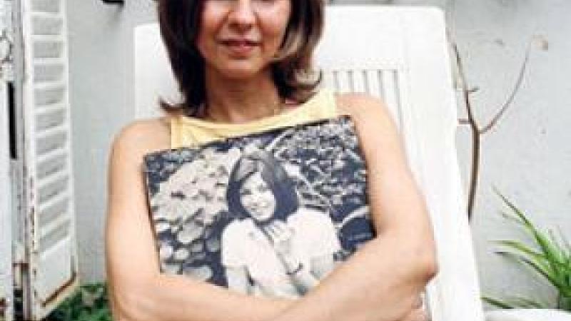 A woman in a sleeveless top sits outdoors on a white lawn chair. She is looking into the camera and her mouth is slightly open in a smile. A black and white image of a smiling young woman rests on her lap, her arms encircling the photograph. In the background are several potted plants and a white window shutter.