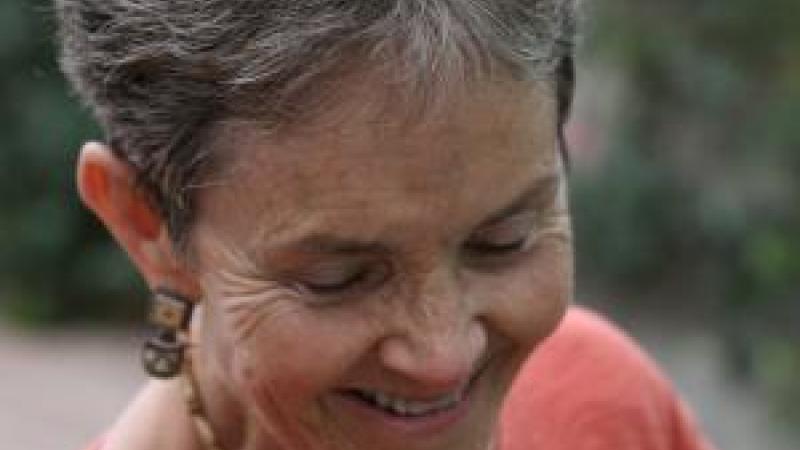 An image from the New Day film States of Grace. Dr. Grace Dammann closes her eyes and smiles towards the ground, her body tilted slightly. Her gray and white hair is short and she wears earrings, a necklace and a long sleeved shirt. The background is full of blurred out foliage.