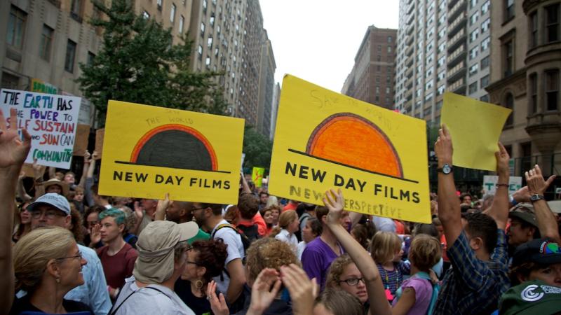 A large group of people of various races, ethnicities and genders mill around in a city street at the People’s Climate March. Several people hold posters up, three of which are yellow films with the New Day Films logo on them. Tall city buildings rise up on either side of the frame.