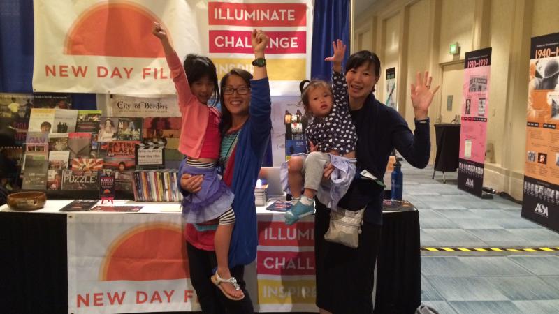 New Day Filmmakers Yun Suh and Debbie Lum each hold a young child on their hip at the New Day Films booth in the exhibition hall. All four smile and raise one fist or open hand above them in celebration.