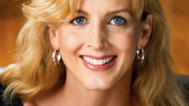 A headshot of New Day filmmaker Kimberly Reed. A trans woman with blonde hair and bright blue eyes smiles widely at the camera wearing hoop earrings. There is a plain orange wall blurred in the background.