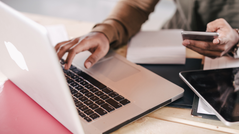 An open laptop on a desk with a hand typing while the other hand is holding a cell phone