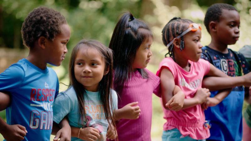 In this still from the film Upstream, Downriver, a group of children are outdoors linking arms.
