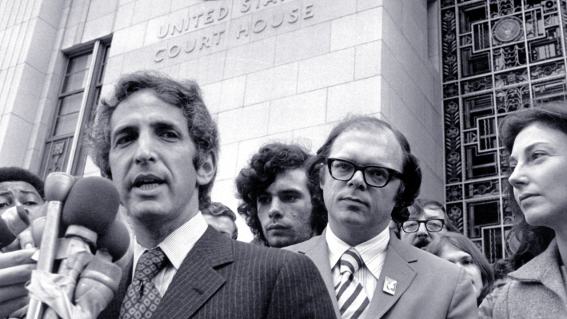 A black and white still image from the film. The camera looks up from below at Daniel Ellsberg, a white man in a pinstriped suit, who speaks into a mass of TV reporter microphones that are taped together. He stands in front of tall, pale building with “United States Court House” embossed on the exterior wall. He is surrounded by men of various ethnicities who listen on or jockey into position to get their microphone closer to him.