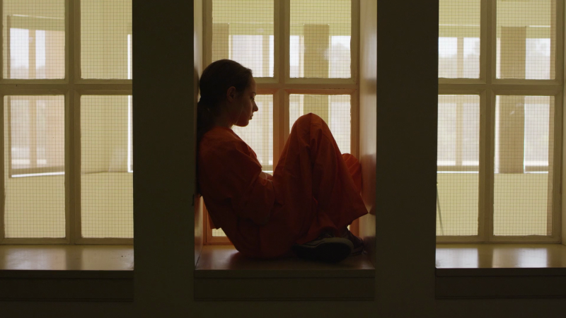 A young woman sits looking out a window in a white room while wearing an orange jumpsuit 