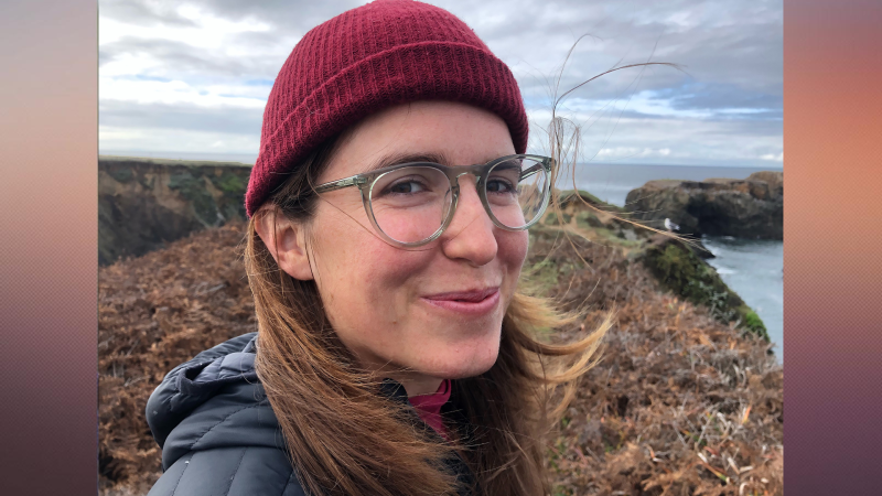 A person with long hair and glasses and wearing a red hat, looks into the camera and smiles. They are outdoors.