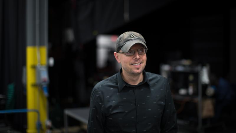 A medium shot of filmmaker Adrian Filmadria, who is wearing a black button down shirt and a baseball cap. He is looking off to the right.