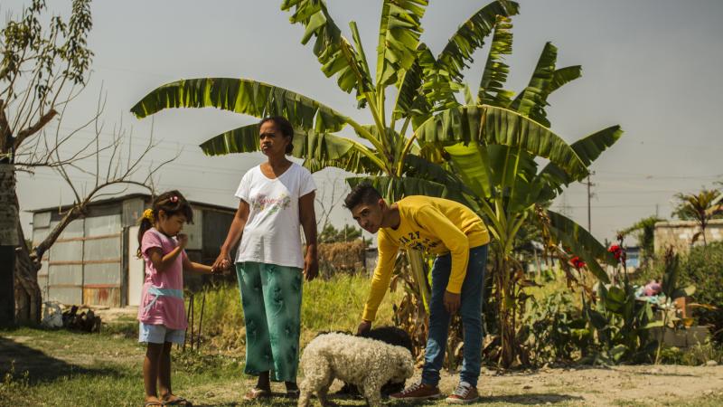 Production Still from Children of Las Brisas. Two adults and a child stand behind a tree in Venezuela. One bends to pet a small dog.