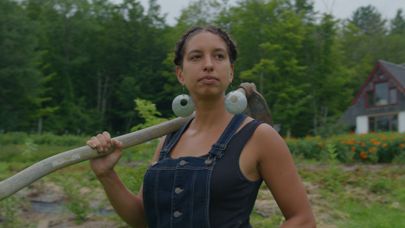 A still from Farming While Black shows a Black woman in overalls stands center with garden hoe over her right shoulder.