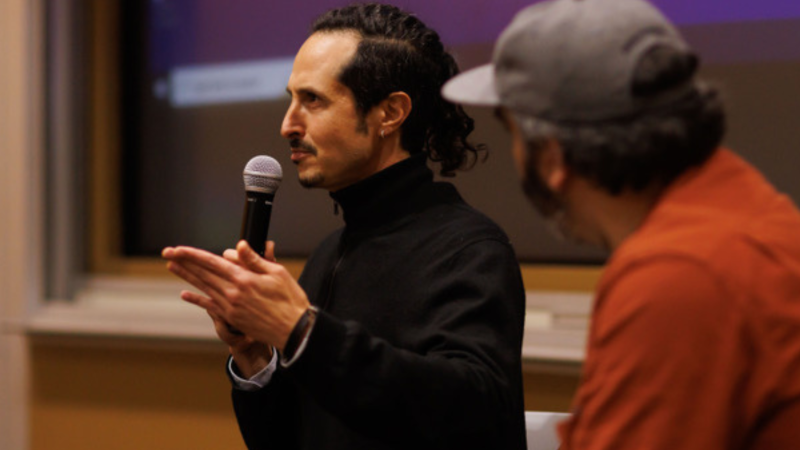 Filmmaker Joel Fendelman, a white man with long, curly black hair, stands at the front of an auditorium addressing an audience. He speaks into a microphone, and another man looks on from beside him.