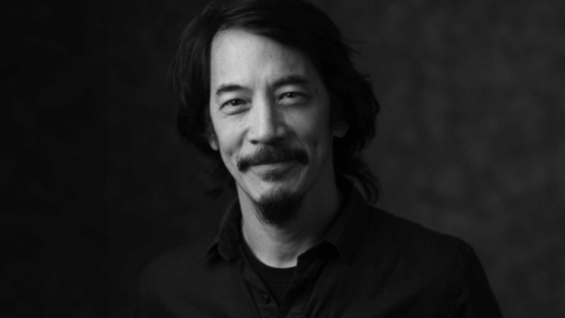 Black-and-white portrait of New Day Films Director Mark Decena. He is smiling at the camera with dark hair, a mustache, and goatee against a dark background.