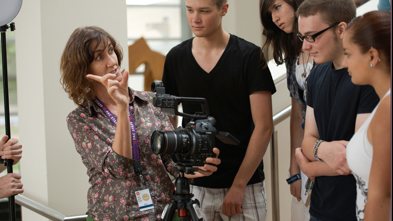  Suzan Beraza, a Latina woman, addresses a group of attentive students around a camera on a tripod. She points to their subject matter off-screen while looking at the students.