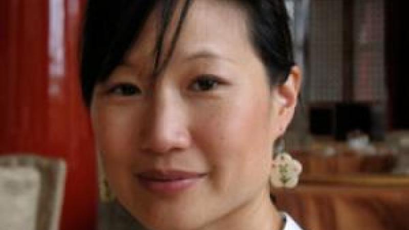 A headshot of filmmaker Debbie Lum smiling softly towards the camera. Her hair is tied back and her side bangs fall across her forehead. She is wearing a white collared shirt, red sunglasses, and flower-shaped earrings.