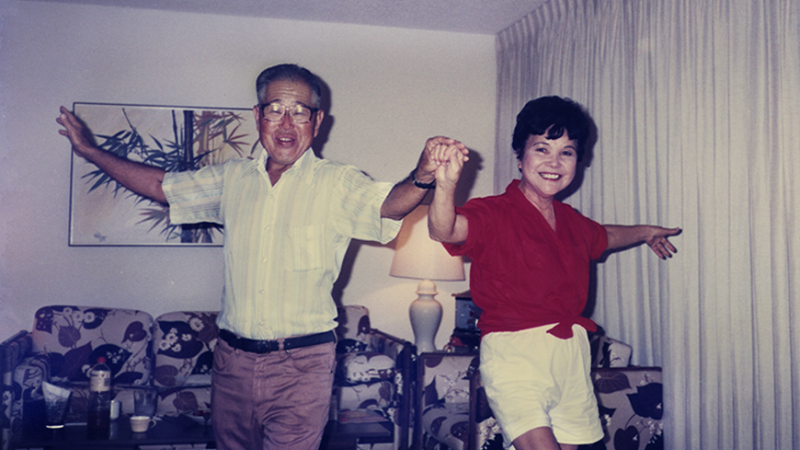 A middle-aged Japanese-American couple hold hands and dance in their living room. Their faces are lit with smiles as they face the camera, arms extended.