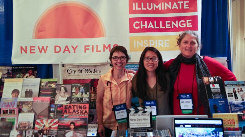 Three New Day filmmakers smile and stand together with conference badges around their necks for the American Sociological Association conference. They’re surrounded by New Day Films including posters, DVDs, a large New Day Films banner, and a laptop with the New Day Films website on it.