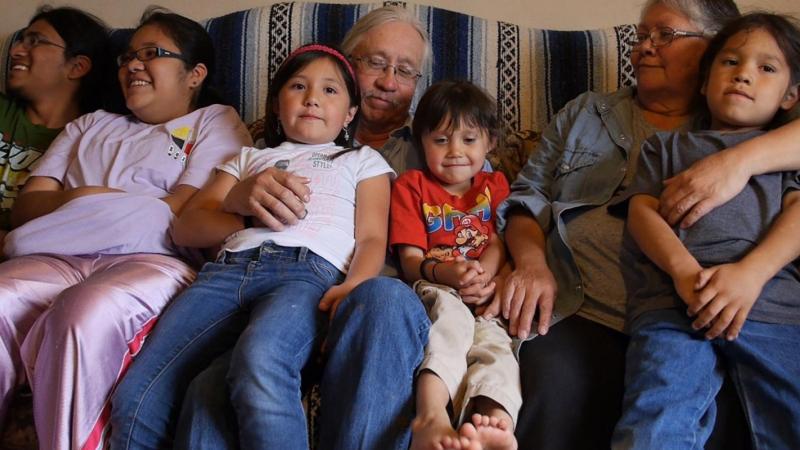 Seven members of a Blackfeet (Pikuni) family, the Mombergs, sit together on a sofa ranging from elders to pre-school age. The youngest three children sit on their grandparents’ laps and look ahead or down while the two older youth smile at something off in the distance.