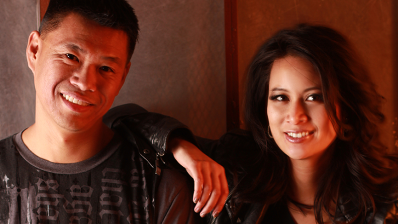 New Day husband and wife filmmaking team Baldwin Chiu and Larissa Lam look directly in the camera smiling softly. Larissa is wearing a leather jacket and has her arm on Baldwin’s shoulder. He is wearing a black shirt with writing on it. They are bathed in red lighting.