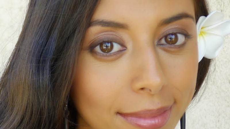 A headshot of New Day filmmaker Chriten Hepuakoa Marquez. A woman with long brown hair and brown eyes wears a white flower tucked behind her ear. She is standing in front of a white background giving a soft, close-mouthed smile to the camera.