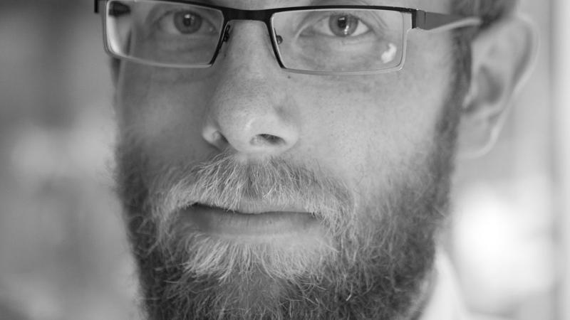 A black and white headshot of New Day filmmaker Christian Jensen. He wears square glasses and a collared shirt and looks into the camera with a serious expression.