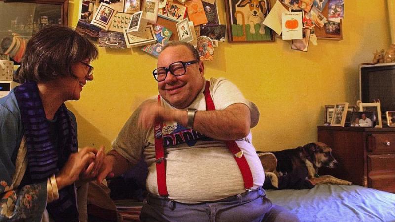 Larry Selman, a community activist and fundraiser with an intellectual disability, and filmmaker Alice Elliot sit, holding hands, talking and laughing in a bedroom. Behind them is a dog, a poster board filled with cards, a dresser with an old TV on top and framed photos.