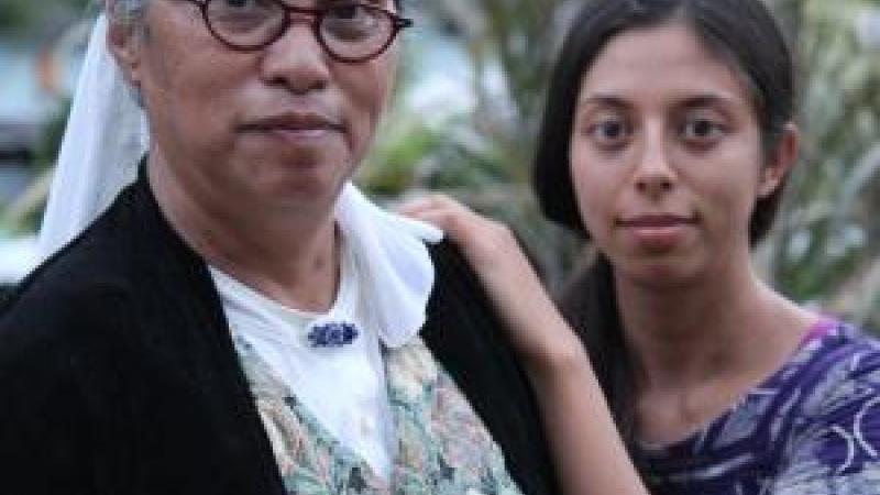 A still from the film E Haku Inoa To Weave a Name. New Day Filmmaker Christen Marquez stands behind her mother and rests her hands on her mother's shoulder. Both women are looking straight into the camera with similar faint smiles on their faces. Lush greenery fills the background.