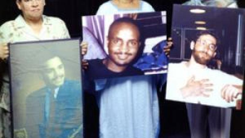 Three women stand shoulder to shoulder and look solemnly at the camera. The each hold a large photo of their late sons.