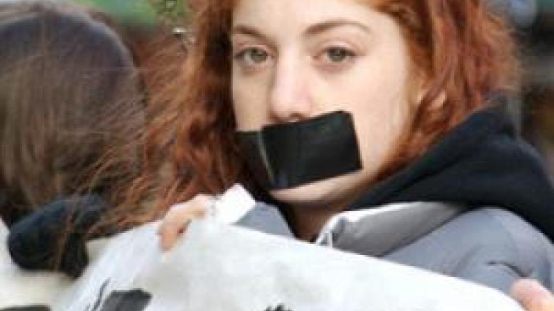 A young girl with red hair stares boldly into the camera in a protest. She has a large black strip of duct tape covering her mouth and she is holding up a protest banner. The other people holding up the banner are blurred in the background.