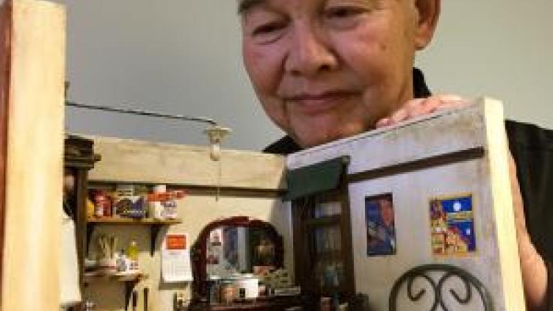 Chinese-American artist Frank Wong peers intently at a detailed diorama that he created of a miniature living room. He points to the inside of the diorama.