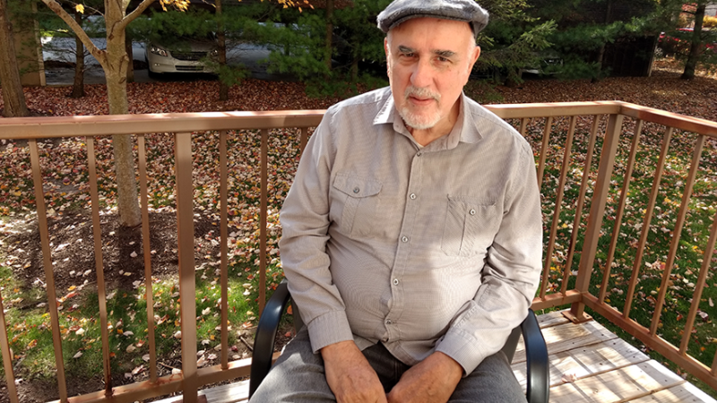 New Day Member George Bogdanich, a white man with a gray goatee, sits on a black chair and looks up at the camera. Behind him is a wooden porch railing and green grass dotted with fallen autumn leaves.