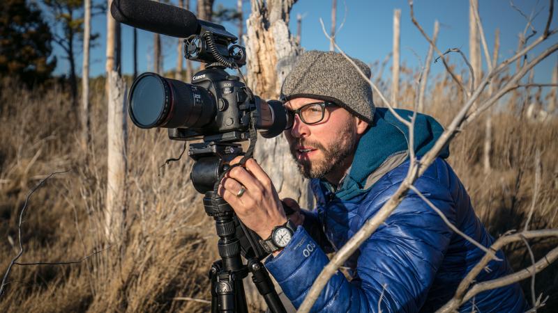 New Day Member Michael O. Synder crouches in a field and looks through a camera that’s mounted on a tripod. The ground is brown, the trees are bare, and Michael wears a blue winter jacket.
