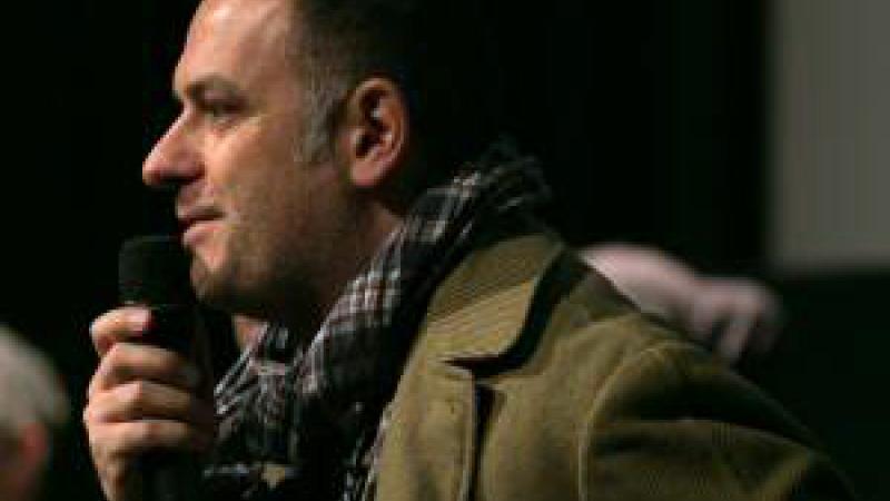A side shot of New Day filmmaker Jean-Michel Dissard. He wears an olive green jacket and a plaid scarf. He holds a microphone to his mouth with an amused expression on his face.