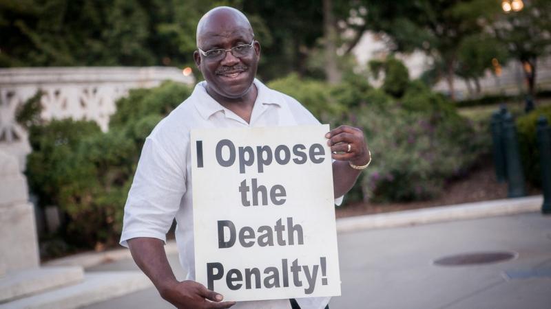Jerry Givens smiles, holding a sign with "I oppose the death penalty!"