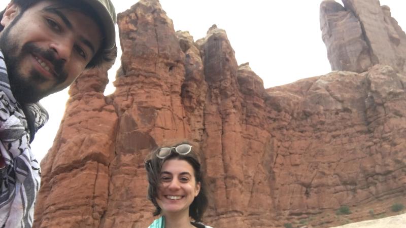 New Day Film directors Jillian Karole and Ahmed Mansour smile at the camera on a windy day. They stand outside with a rocky, sandy cliff towering behind them.